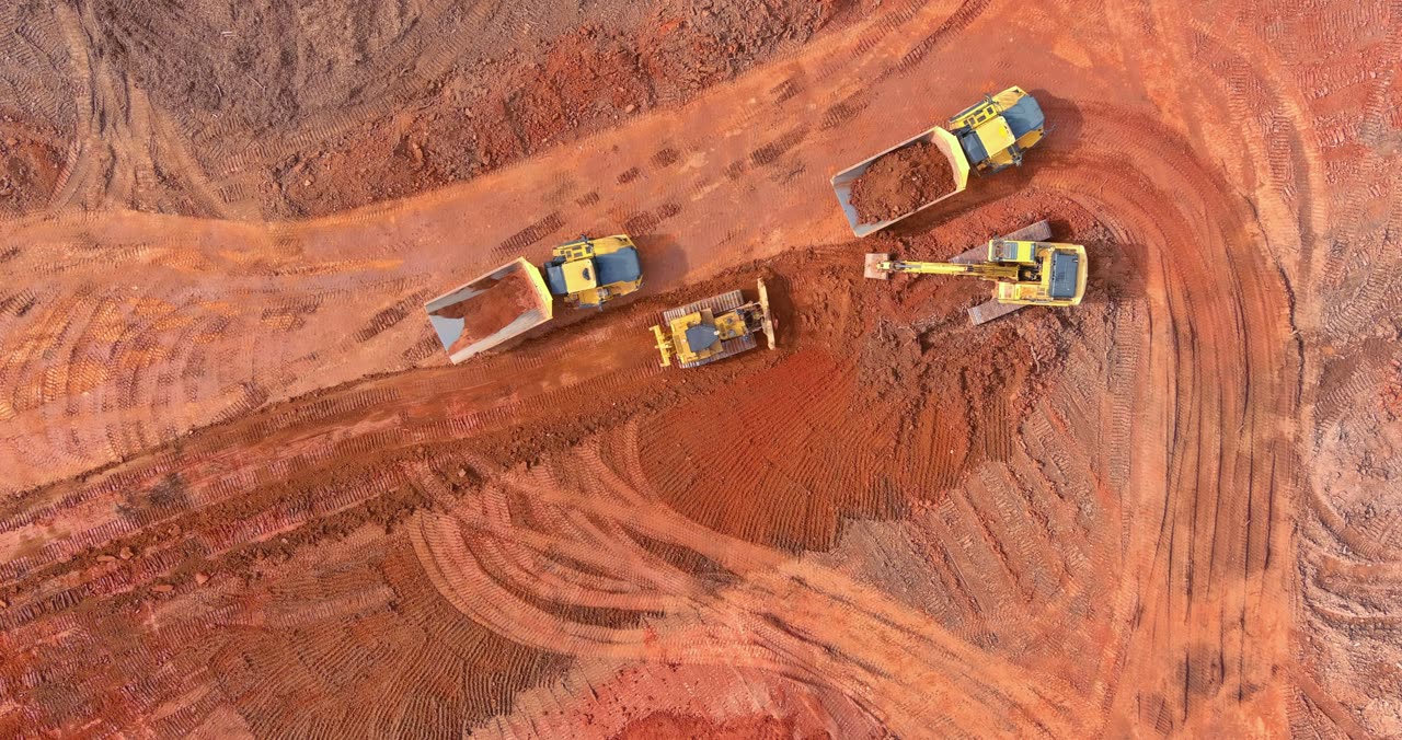 An excavator, a bulldozer and two dump trucks moving red clay, seen from above